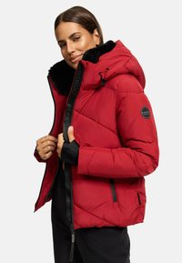 Veste rembourrée rouge avec capuche en fausse fourrure noire et fermeture éclair à l'avant. Elle présente des détails de couture diagonale et un patch logo sur la manche gauche.