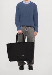 Czarna torba na ramię z podwójnymi uchwytami, z logo na przodzie. Włóknista faktura, pojemny design, trzymana przez osobę w niebieskim swetrze.