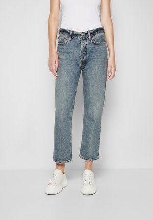 Person trägt blaue Jeans mit geradem Bein und kurzem Schnitt, weißes ärmelloses Oberteil und weiße Turnschuhe, steht vor einem einfarbigen hellen Hintergrund.