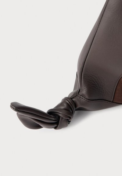 Gros plan d'un sac en cuir marron foncé avec un détail de nœud sur la sangle, sur un fond clair uni.
