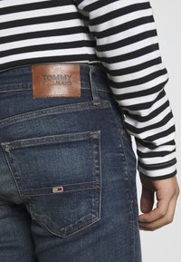 Tmavě modré džínové kalhoty s koženou nášivkou TOMMY JEANS na pásku. Mají zadní kapsy a malý logotyp v červené, bílé a modré barvě.