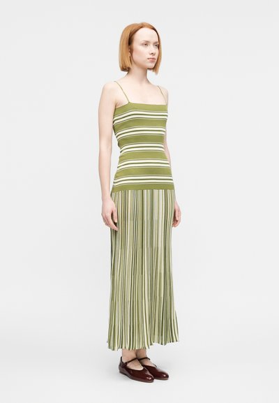 Γυναίκα με μακριά φόρεμα χωρίς μανίκ ια, ριγέ σε ελαιώδη πράσινη και λευκή απόχρωση με λεπτές τιράντες, φοράει σκοτεινά καφέ παπούτσια Mary Jane, στέκεται μπροστά σε λευκό φόντο.