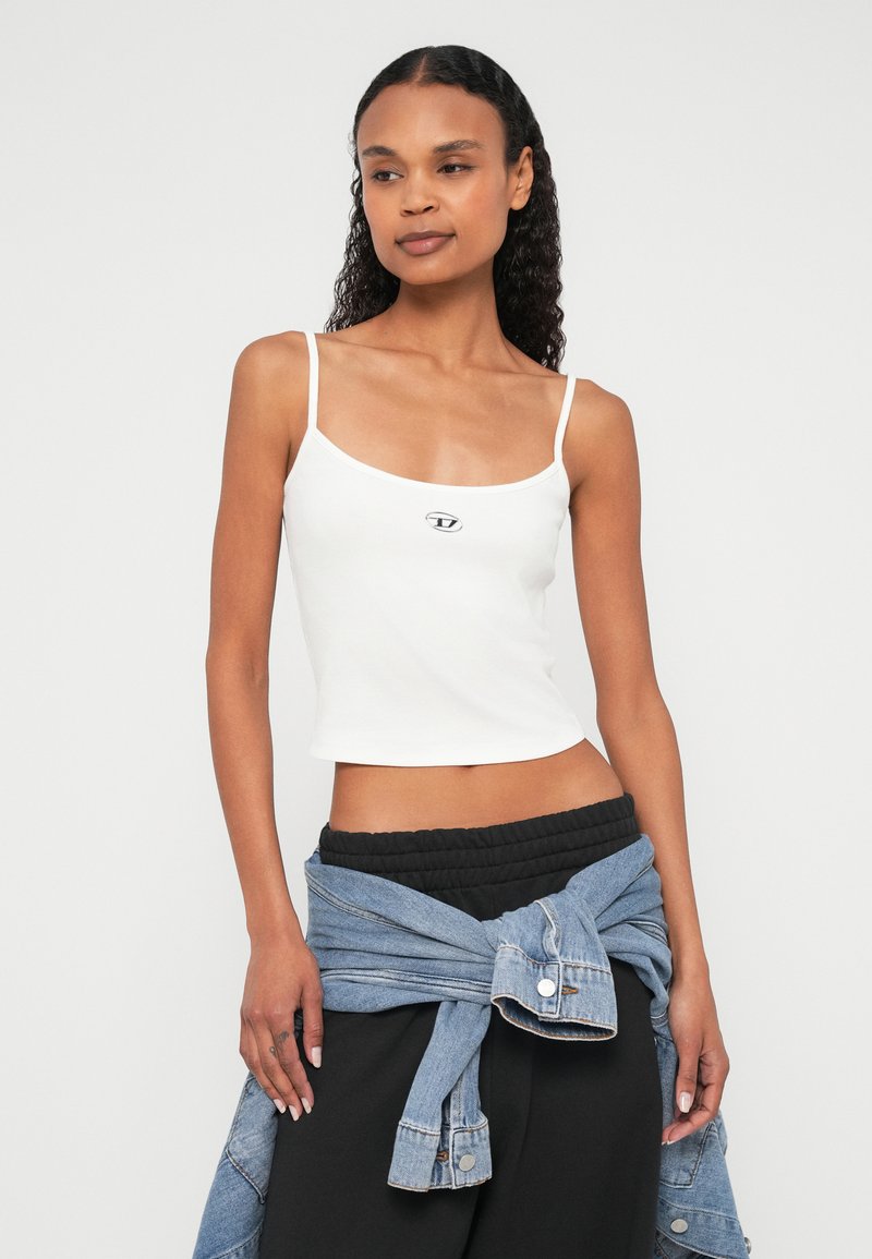 Top corto blanco con tirantes finos, con un pequeño logo negro en el centro. Modelo con pantalones negros y chaqueta vaquera anudada a la cintura.