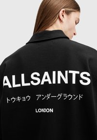 Musta huppari, jossa on kaulus. Selässä suurilla valkoisilla kirjaimilla lukee "ALLSAINTS" ja muita merkkejä. Sileä kankaan rakenne.