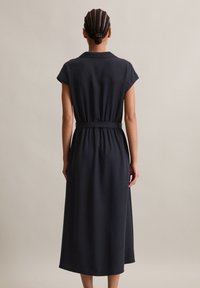 Frau mit geflochtenem Haar trägt ein schwarzes, kurzärmeliges Midikleid mit einem Gürtel und steht vor einem schlichten hellen Hintergrund, wobei sie sich rückwärts wendet.