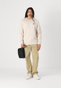 Beige genser med ribbede mansjetter og hem, kombinert med lysebeige bukser og hvite joggesko. Holder en svart veske. Enkel, uformell design.