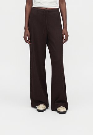 Persoon die losse donkerbruine broek met wijde pijpen en een koord in de taille draagt, en beige schoenen, staande tegen een effen achtergrond.