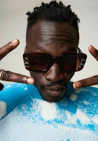 Zonnebril met een gedurfde rechthoekige vorm en donkere lenzen; gemuste bruin en crèmekleurige steunen. Draagt een blauw-wit gemusterde trui.