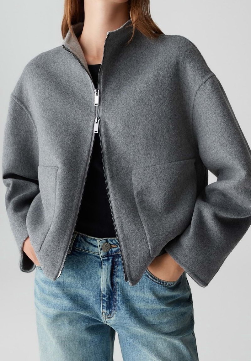 Personne portant une veste grise zippée avec de grandes poches par-dessus un haut noir et un jean bleu, les mains dans les poches de la veste.