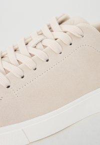 Beige semsket sneakers med flate lisser, lett tekstur og en hvit gummisåle. Synlig søm-detalj langs sidene.