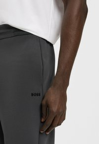 Mörkgrå byxor med en slät textur som har en diskret präglad "BOSS"-logotyp på sidan, matchat med en vit skjorta.