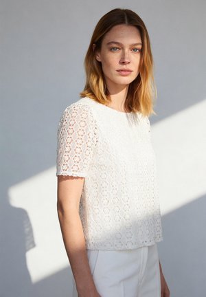 Young woman with shoulder-length hair wears a white lace short-sleeve top and white pants, standing against a light gray wall.