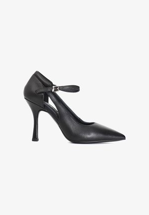 Zapato de tacón alto de cuero negro con puntera puntiaguda, correa en el tobillo y hebilla plateada, tacón stiletto delgado, perfil lateral sobre fondo blanco.