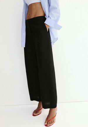 Personne portant un pantalon noir ample, une chemise rayée bleu clair et des tongs rouges, debout, les mains dans les poches, sur fond blanc.