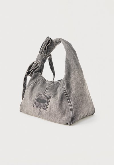Χιαστί τσάντα ώμου από γκρι τζιν με φαρδιά τιράντα δεμένη σε φιόγκο και μικρή διακριτική λεπτομέρεια στο εμπρόσθιο μέρος, σε μονόχρωμο φόντο.