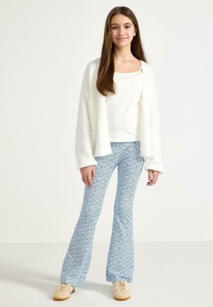 Nuori tyttö seisoo valkoinen toppi, pörröinen valkoinen neuletakki, siniset kukkakuosiset leveälahkeiset housut ja beiget lenkkarit päällä, hymyillen kameralle.