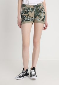 Maskovací šortky v zelené a béžové barvě s listovým vzorem, vybavené pěti kapsami a poutky na opasek, spárované s černými vyššími teniskami.