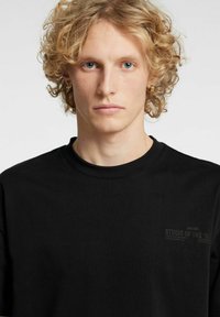 Svart bomulls-T-skjorte med rund hals, korte ermer og subtilt trykt tekstdetalj på venstre brystområde. Jevn tekstur.