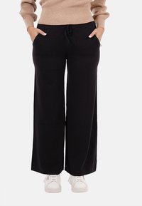 Persona che indossa pantaloni neri a gamba larga con coulisse e un maglione beige, con le mani in tasca; scarpe da ginnastica bianche visibili sotto i pantaloni.