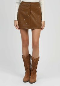 Mini-jupe en cuir marron avec des clous en argent, associée à des bottes de cow-boy en daim beige et un haut blanc à manches longues, visible de la taille vers le bas.