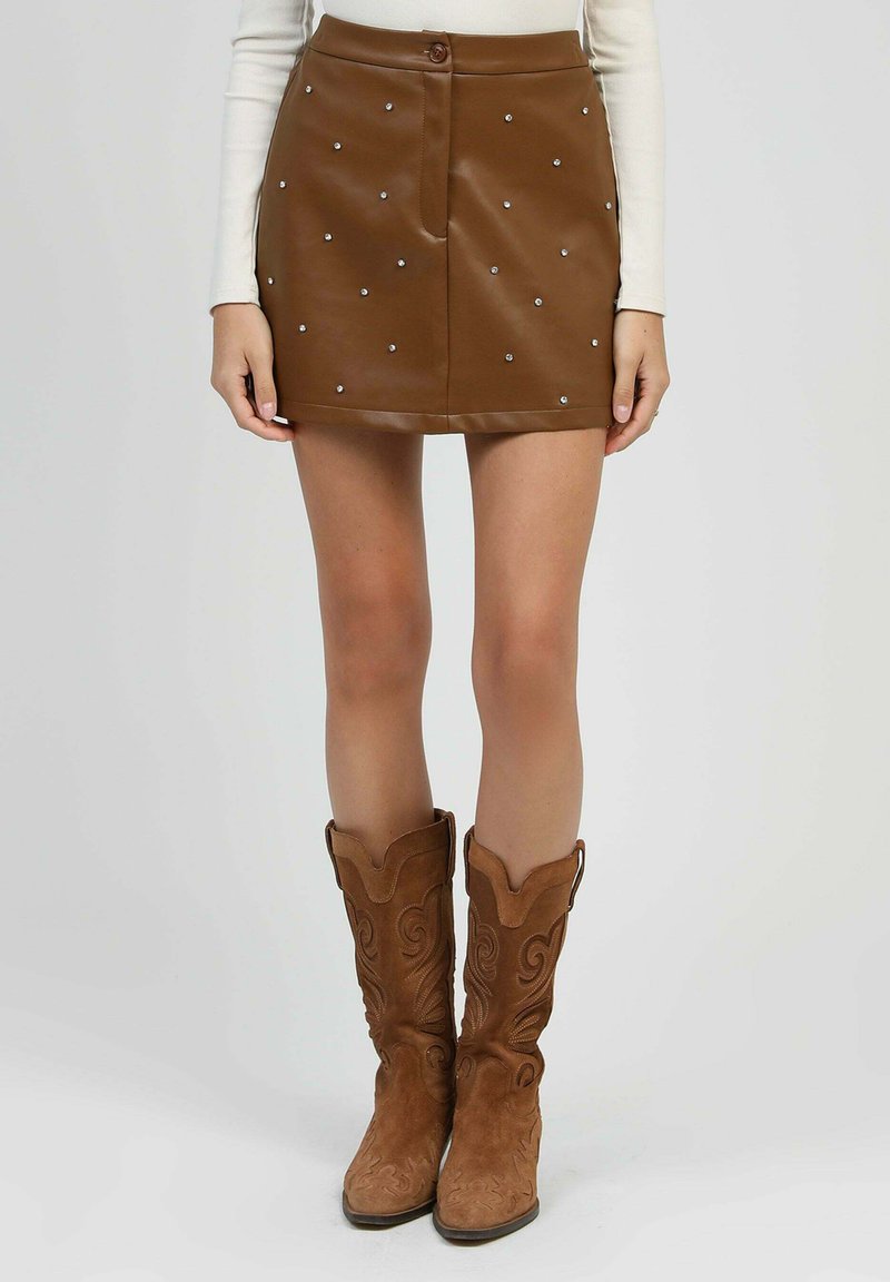 Mini-jupe en cuir marron avec des clous en argent, associée à des bottes de cow-boy en daim beige et un haut blanc à manches longues, visible de la taille vers le bas.