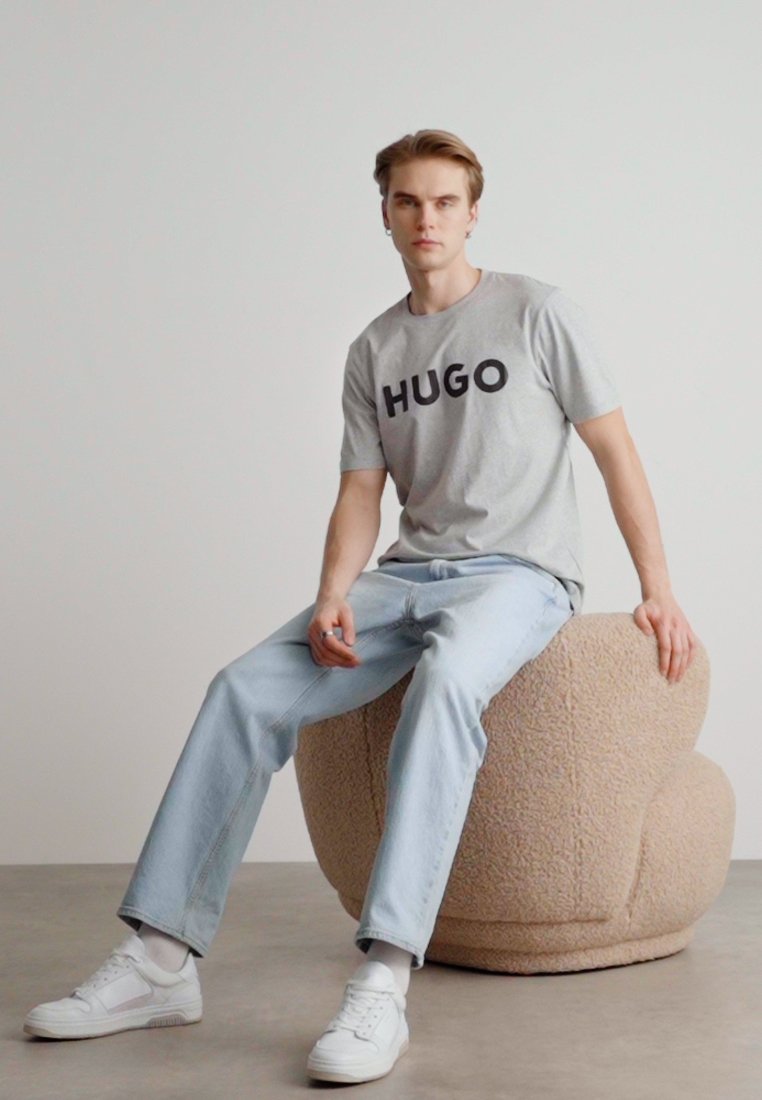 Сив T-shirt с текст "HUGO" в черно, светлосини дънки, бели маратонки, седнал на бежов, текстуриран кръгъл табуретка, минималистичен фон.