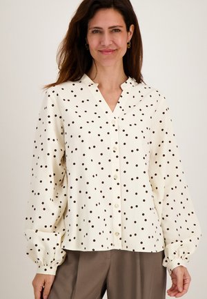 Vrouw draagt een witte blouse met zwarte stippen en lange pofmouwen, gecombineerd met losse bruine broek.