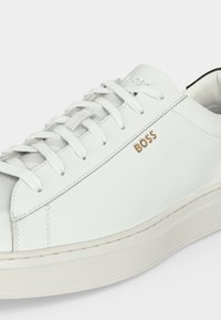 Baskets en cuir blanc avec lacets blancs, logo beige "BOSS" sur le côté, et semelle épaisse blanc cassé, inclinée pour montrer le dessus et le côté extérieur.