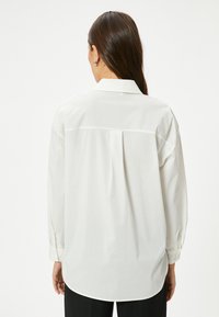 Camicia bianca a maniche lunghe con colletto classico, dotata di un dettaglio plissettato sul retro e polsini con bottoni, realizzata in un tessuto morbido e liscio.