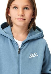 Ljusblå zip-up huvtröja med en framficka, med en logotypdesign i vitt. Tillverkad av mjukt tyg, med dragsko på huvan och detaljer i blixtlås.