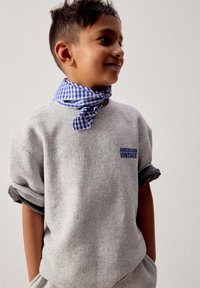 Junge trägt einen grauen Pullover mit dem Schriftzug "American Vintage" und einen blau karierten Schal um den Hals gebunden, Hände in den Taschen, lächelnd.