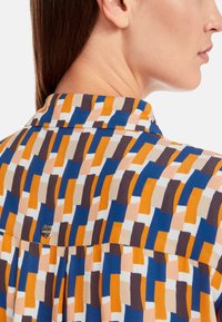 Bunte, gemusterte Bluse mit rechteckigen Formen in Blau, Orange, Braun und Creme. Verfügt über einen Kragen und ein metallisches Logo-Detail auf der Rückseite.