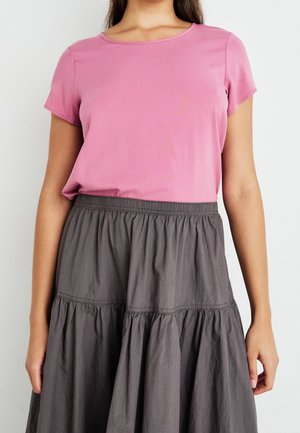 Woman wearing a short-sleeve pink blouse tucked into a high-waist, gathered dark gray skirt, standing against a plain white background.