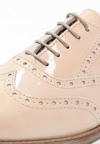 Chaussure en cuir beige avec finitions brillantes, détails perforés élaborés, lacets gris clair et texture lisse. Bout rond et design épuré.