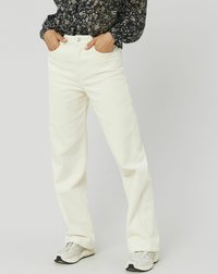 Personne portant un pantalon droit blanc cassé, un blouse à manches longues à motifs floraux et des baskets de couleur claire, les mains dans les poches, debout devant un fond uni.