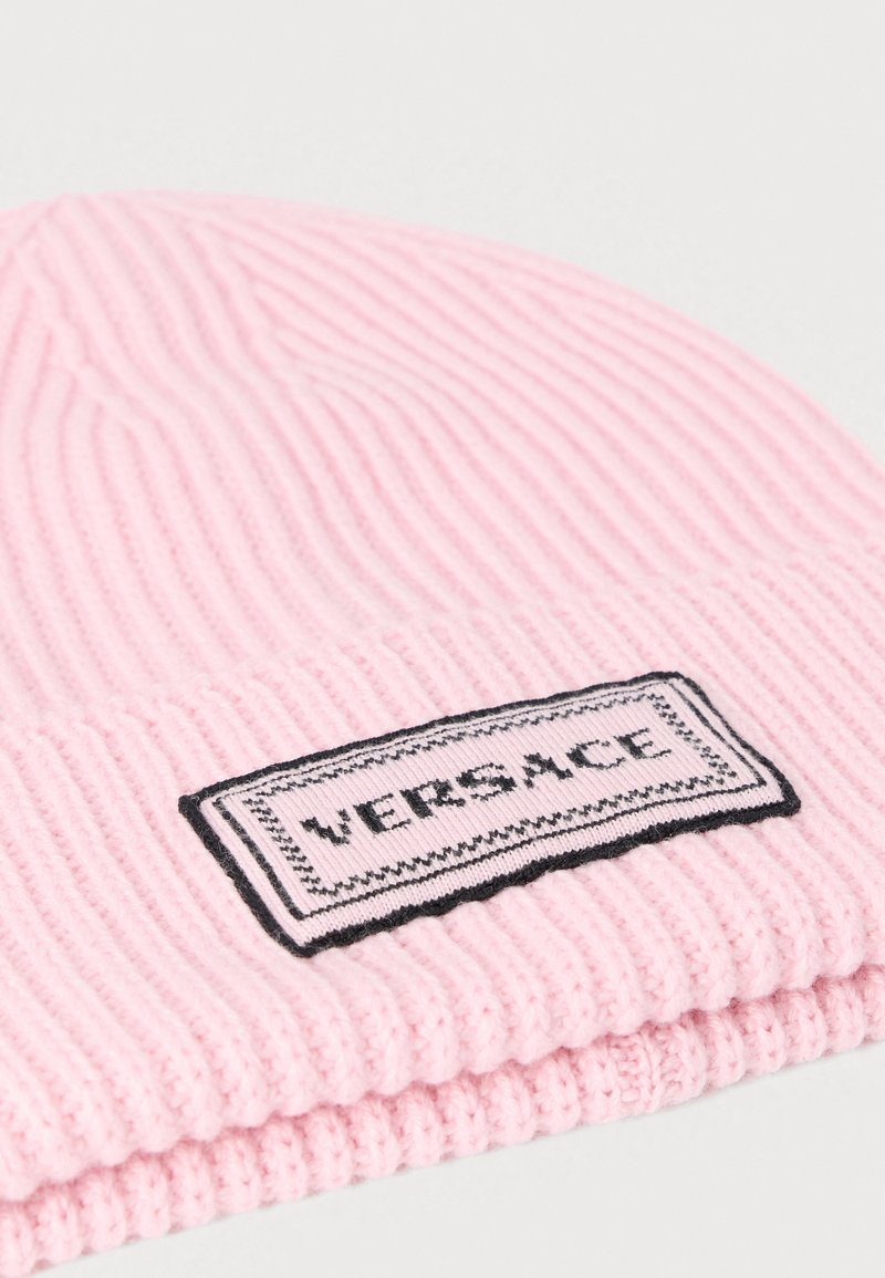 Pink ribbed knit beanie with a rectangular black and white label reading "VERSACE." Soft texture with folded edge.