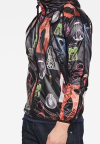 Veste légère à capuche avec un fond noir orné d'un collage de motifs photographiques colorés représentant divers objets et outils.