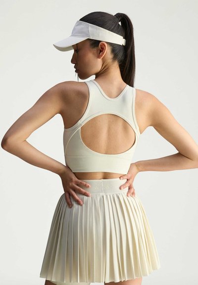 Sujetador deportivo beige con un diseño de espalda en forma de scoop, combinado con una falda plisada a juego. Ambos hechos de una tela suave y texturada. Visera blanca en la cabeza.