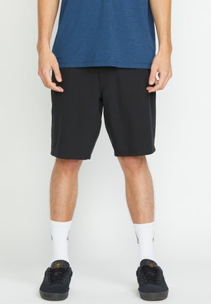 Personne portant un short noir, des chaussettes blanches montantes, des baskets noires et un t-shirt bleu, debout sur un fond blanc.