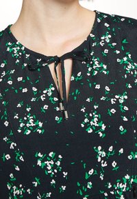 Vestido negro con estampado floral en blanco y verde, que presenta un escote en forma de ojo de llave y un cierre tipo lazo con puntas metálicas.