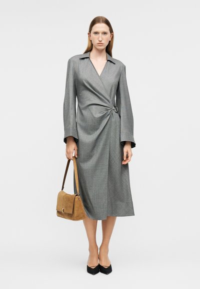 Γυναίκα με μακριά γκρι φόρεμα τύπου wrap, κρατώντας μια τσάντα χειρός από ταμπά σουέντ και φορόντας μαύρες μυτερές γόβες, που στέκεται μπροστά σε απλό φόντο.