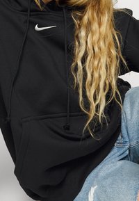 Svart Nike-hoodie med framficka, dragsko i huvan och en vit swoosh-logga. Kombinerad med ljusblå slitna jeans.