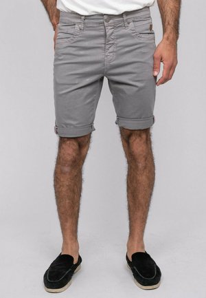 Homme portant un short gris retroussé, debout les mains partiellement dans les poches, avec des chaussures noires sans lacets sur un fond uni.