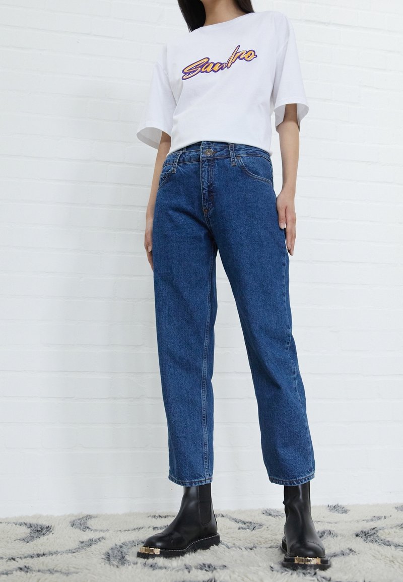 Personne portant une chemise blanche à manches courtes avec le texte "Sandro", un jean bleu taille haute et des bottines noires, debout sur un tapis à motifs.
