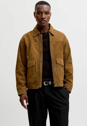 Young man with short hair wearing a brown suede jacket over a black sweater and black pants, standing against a white background.