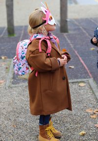 Bruine wollen jas, roze rugzak met vogelpatroon, gele laarzen en een fantasierijk eenhoornmasker. Kind staat zijwaarts in een parkomgeving.