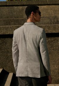 Blazer gris clair avec épaules structurées, texture de tissu naturel, fente centrale au dos et accents de boutons subtils sur les poignets.