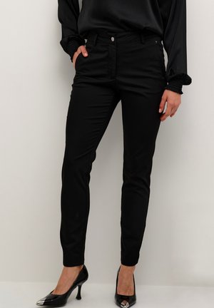 Black fitted pants made of stretch fabric, featuring a zip fly, five-pocket design, and a smooth texture, paired with shiny black heels.