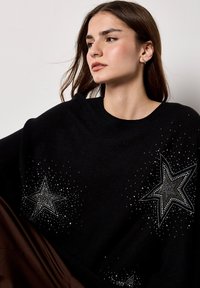 Czarny sweter dzianinowy z wzorem gwiazd, ozdobiony cyrkoniami, prezentujący luźny krój i szeroki dekolt. Materiał wydaje się miękki i teksturowany.