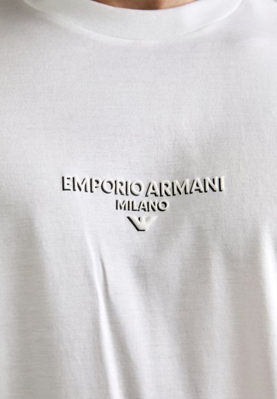 White t-shirt with embossed black text reading "EMPORIO ARMANI MILANO" and a stylized eagle logo below the text.